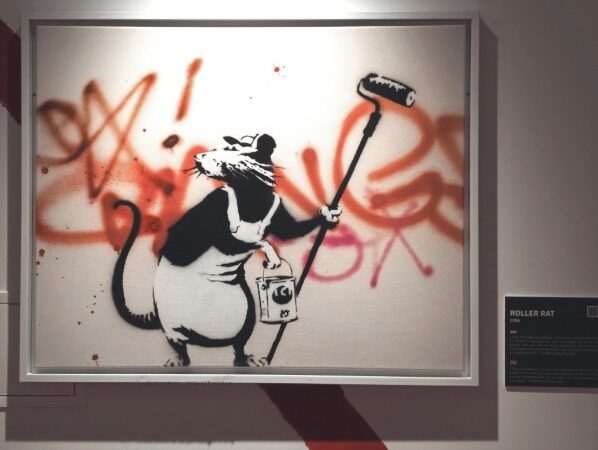 Banksy-style rat holding a paint roller in front of graffiti background inside a frame
