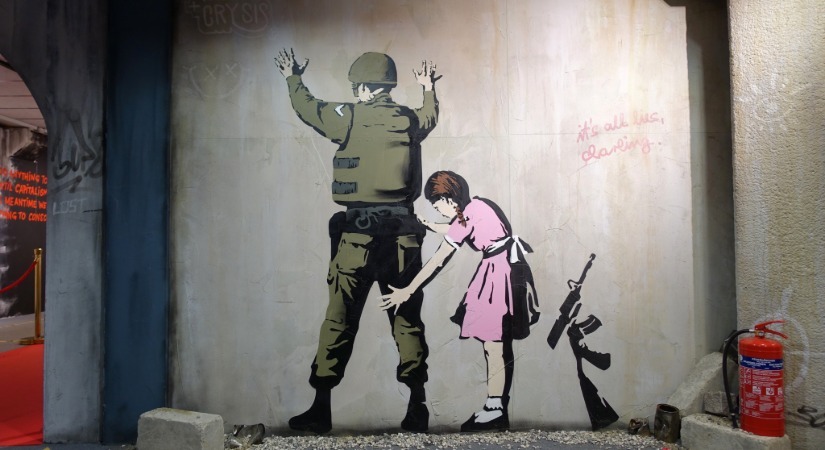 Banksy-style mural of a young girl frisking a soldier against a wall