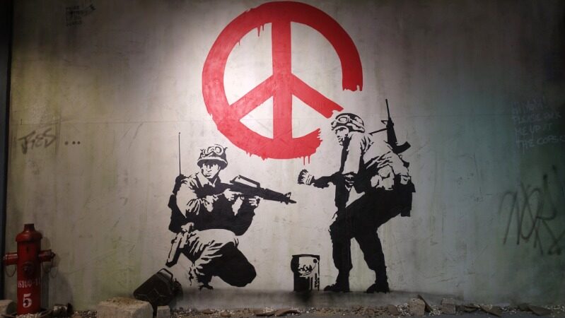 Banksy-style mural of two soldiers painting a red peace symbol on a wall